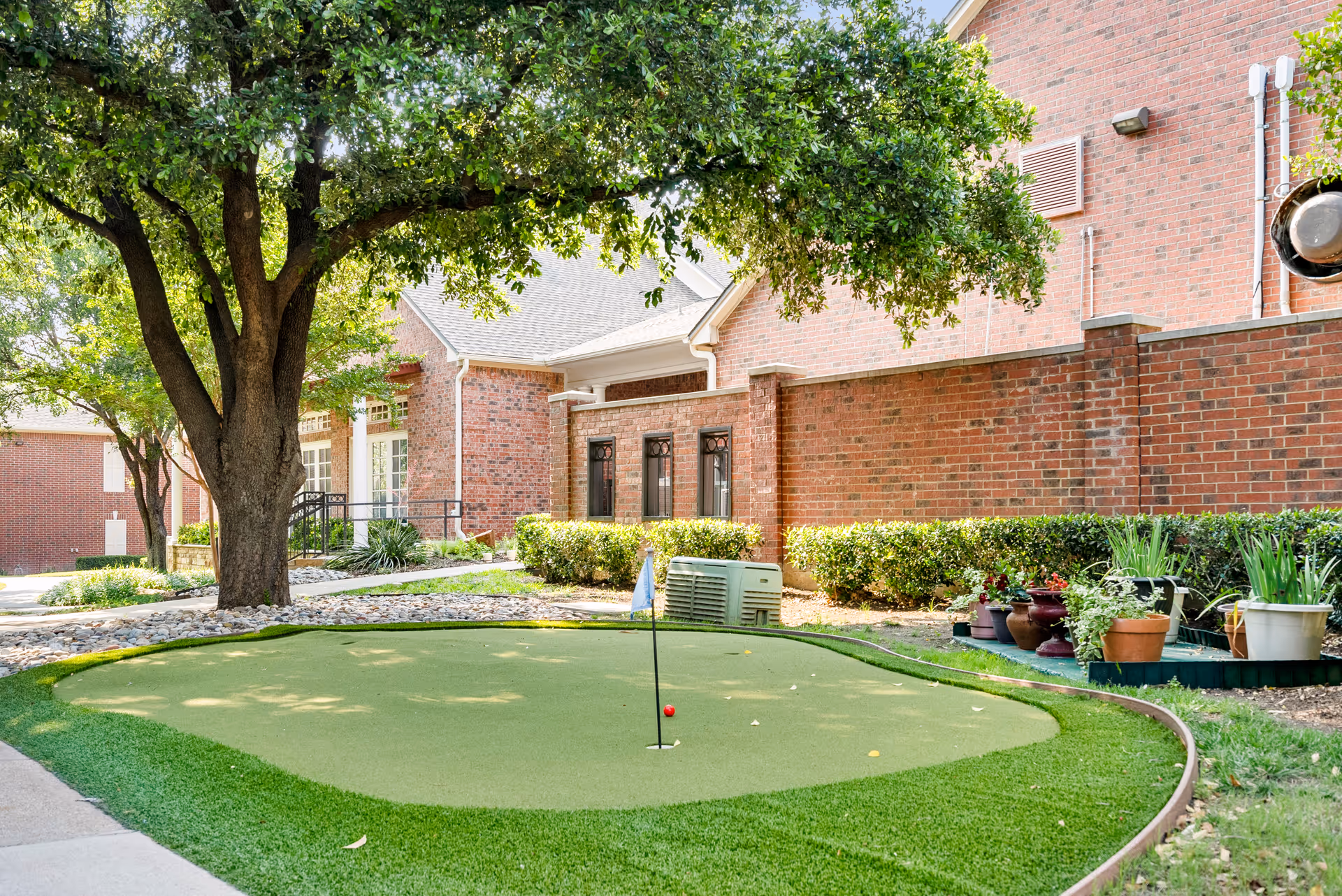 Outdoor putting green with a small flag and a red golf ball, surrounded by green grass, potted plants, and a large tree. In the background, there is a brick building with windows and shrubs along the wall.