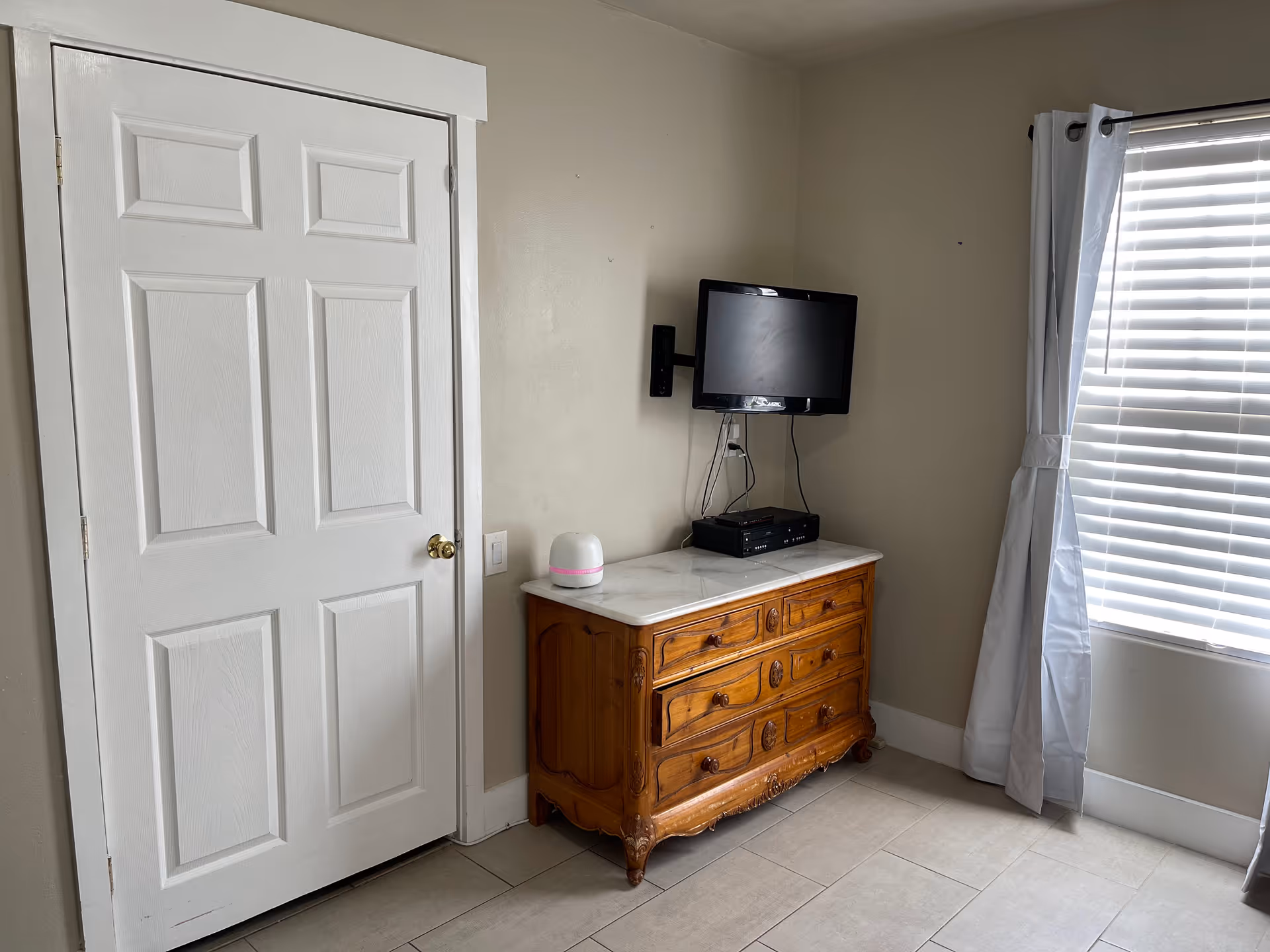 A corner of a room with a closed white door on the left, a wooden dresser with a marble top in the center, and a small flat-screen TV mounted on the wall above the dresser. A white device with a pink stripe sits on the dresser. To the right is a window with white blinds and light gray curtains tied back.