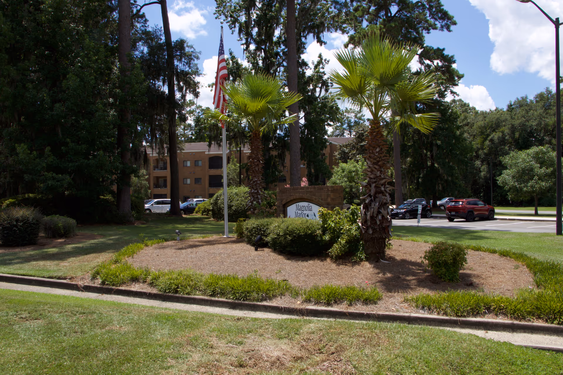 Outdoor view of Magnolia Manor of Richmond Hill with a landscaped area featuring palm trees, bushes, an American flag on a flagpole, and a sign partially visible behind the greenery. In the background, there is a multi-story building and a parking lot with several cars under a partly cloudy sky.