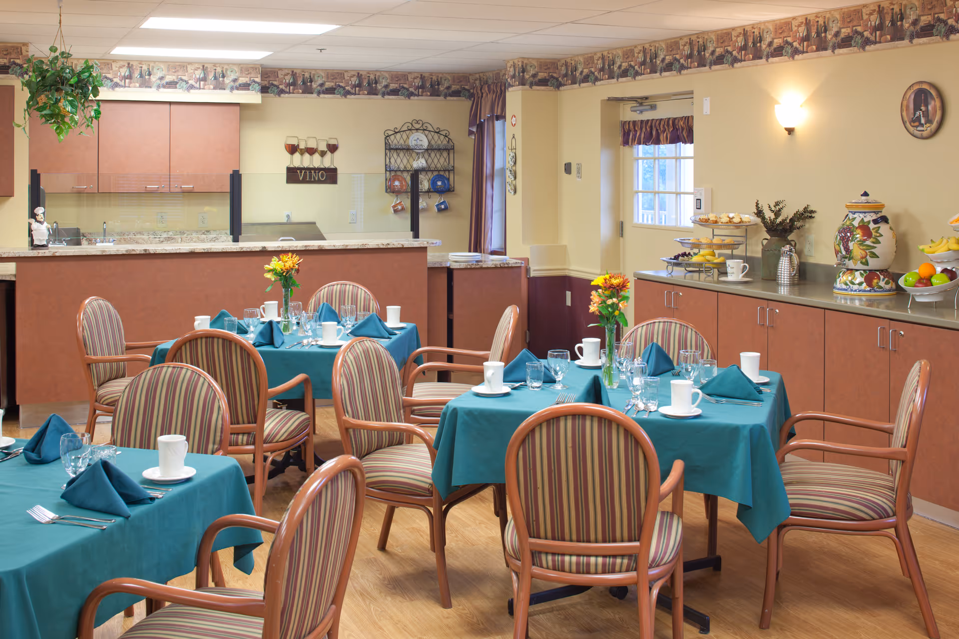 A dining room with several tables covered in teal tablecloths, each set with white cups, glasses, silverware, and folded teal napkins. The chairs have wooden frames and striped upholstery. In the background, there is a kitchen area with cabinets and a counter, along with decorative items such as a wine glass wall decoration, a fruit bowl, and a large decorative ceramic container. The room has warm lighting and a cozy atmosphere.