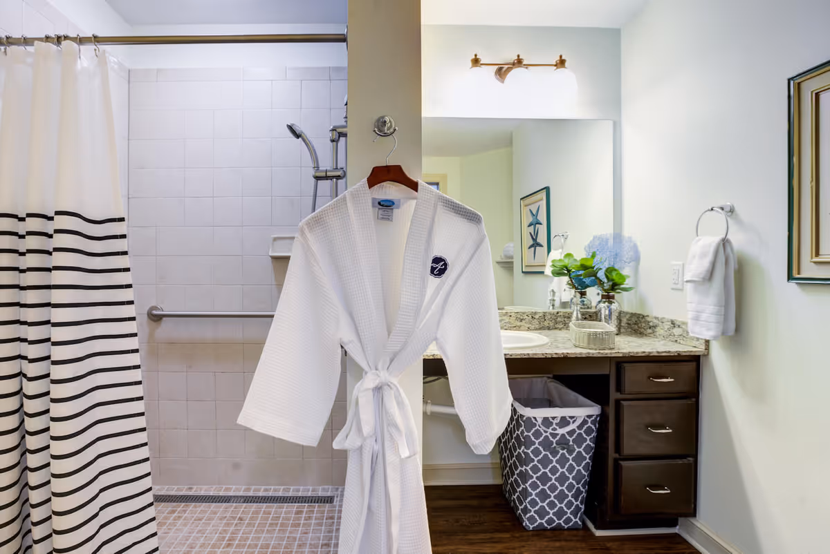 A bright bathroom showing a white robe hanging on a hook in front of a tiled shower, a vanity with sink and mirror, and a patterned laundry basket.