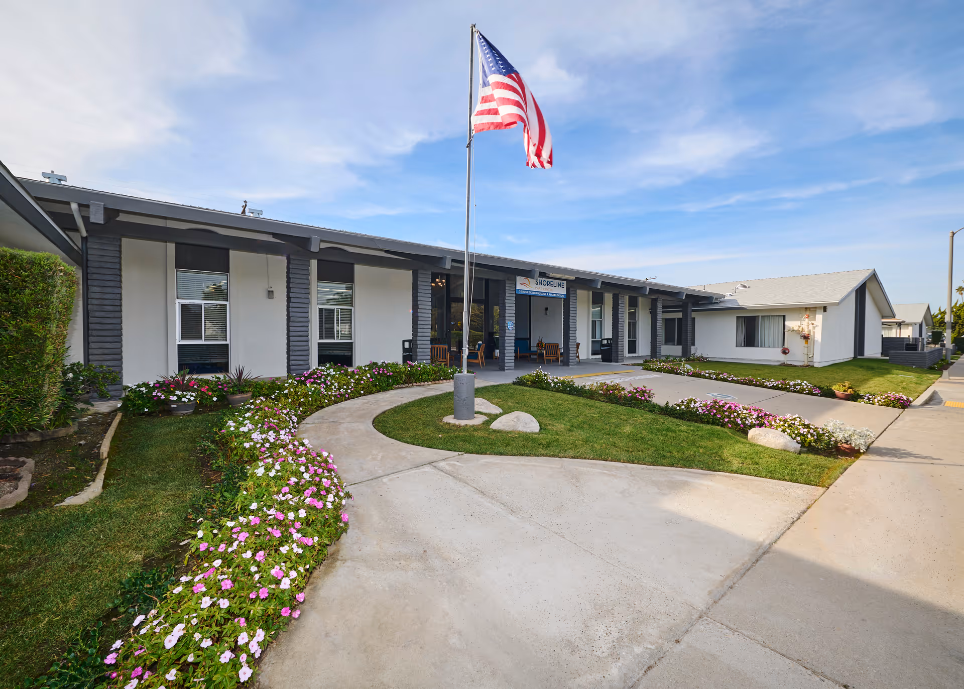 Exterior view of Shoreline Care Center building with a curved walkway, green lawn, flower beds, and an American flag on a flagpole in front of the entrance under a partly cloudy sky.