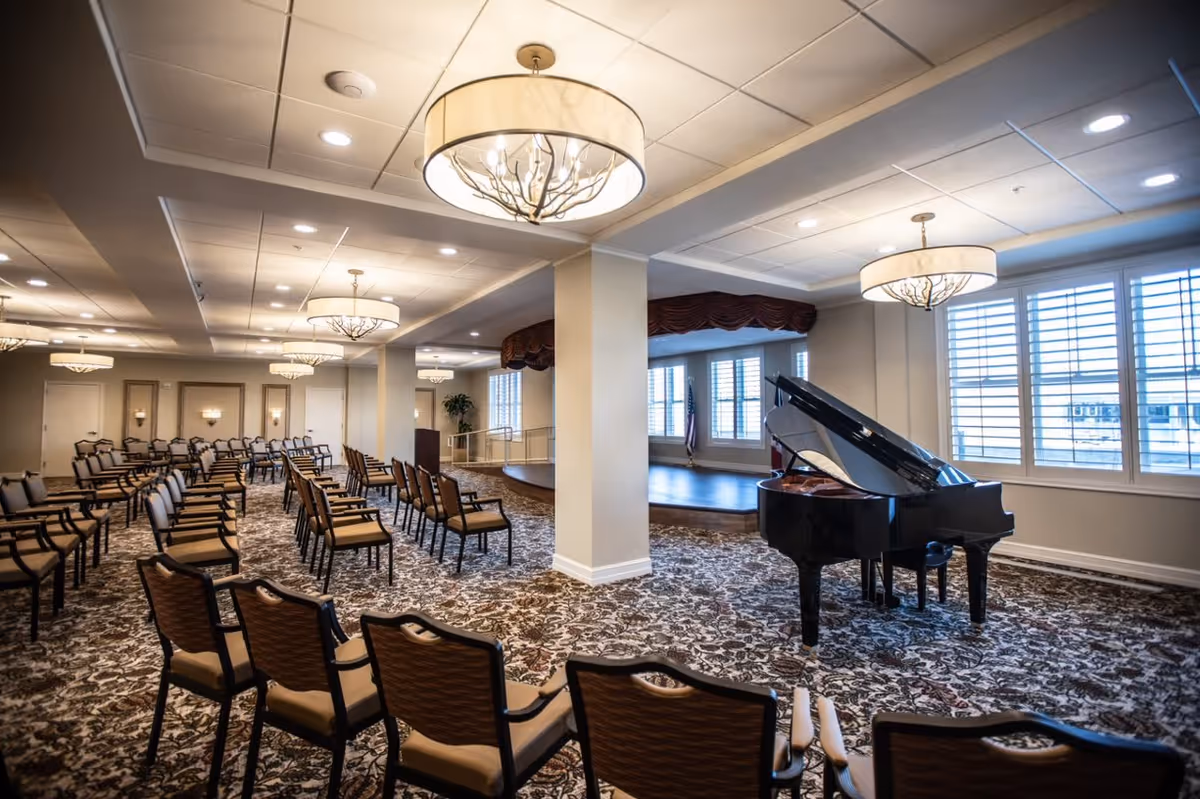 A spacious event room with rows of chairs arranged facing a small stage area. A black grand piano is positioned near the stage, and large windows with white shutters allow natural light to fill the room. The ceiling features multiple decorative light fixtures, and the floor is covered with a patterned carpet.