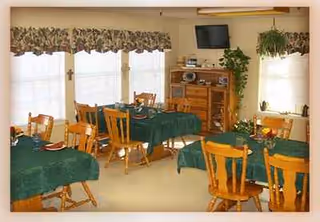 Casual dining room with green tablecloths, wooden chairs, windows, and a cabinet with plants.