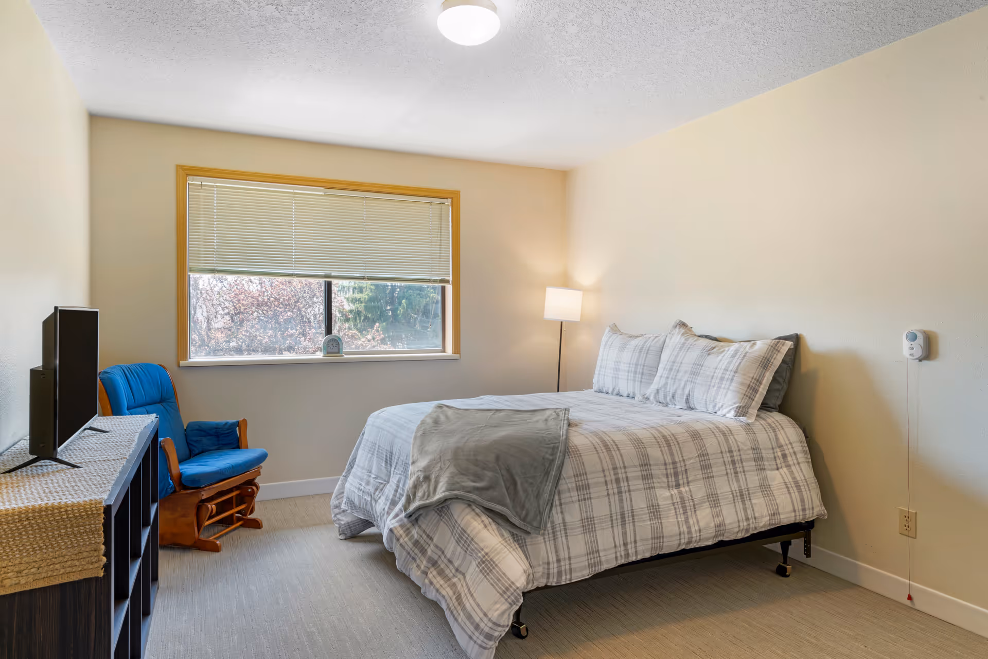 A bedroom with a bed made with plaid bedding and a gray throw blanket. There is a blue cushioned wooden rocking chair next to a window with partially closed blinds. A floor lamp is positioned in the corner near the bed, and a TV sits on a dark wooden shelf covered with a beige runner.
