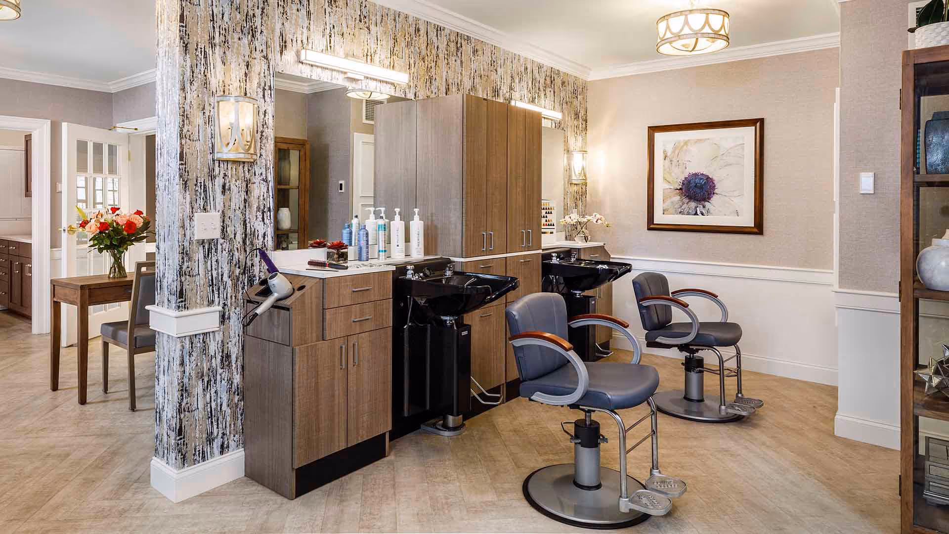 Interior view of a salon area in a senior living facility with two black shampoo sinks and two gray salon chairs with wooden armrests. The space features wood cabinetry, a large mirror, wall sconces, and a framed floral artwork on the wall. In the background, a dining area with a table and chairs and a vase of flowers is visible.