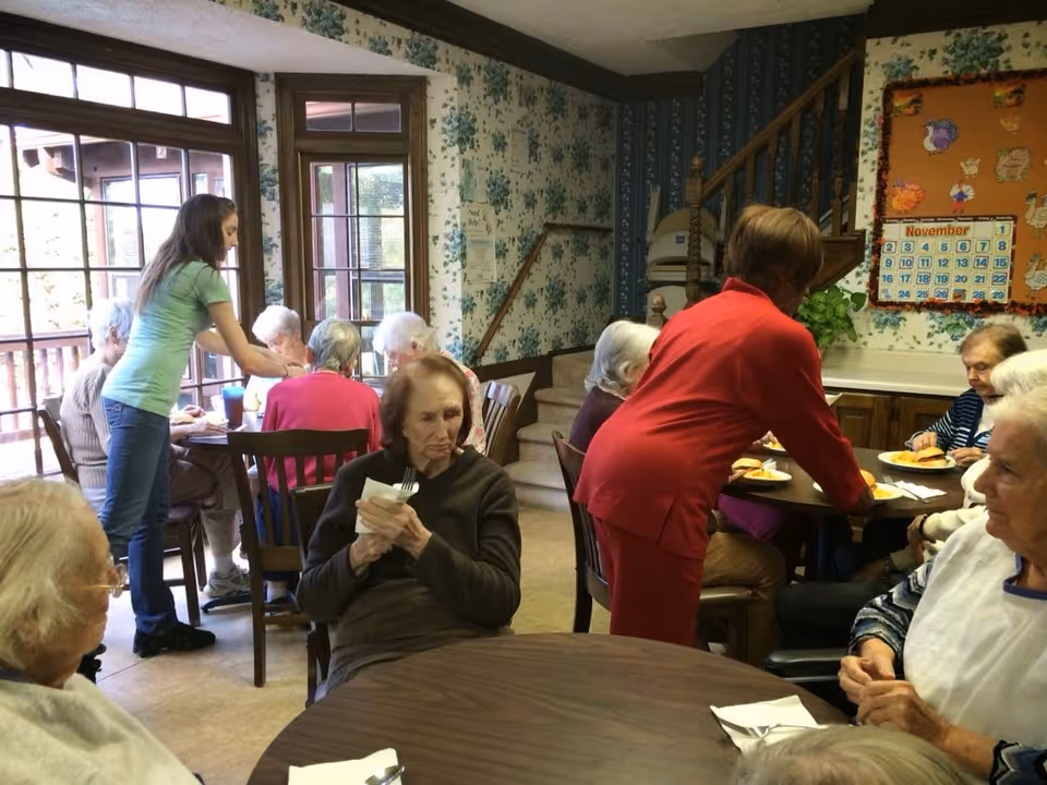 A group of elderly people seated around tables in a dining area, being assisted by two caregivers. The room has floral wallpaper, large windows, and a bulletin board with a November calendar on the wall.