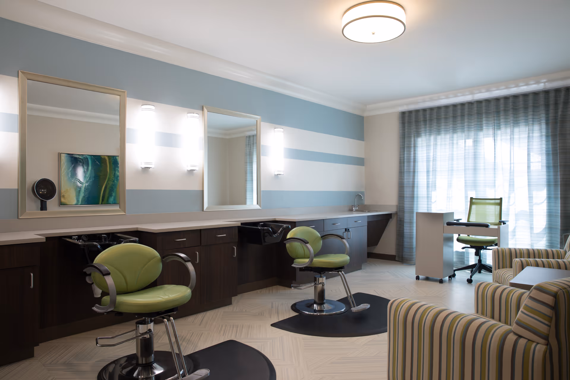 A bright and modern salon area in a senior living facility featuring two green salon chairs in front of mirrors with wall-mounted lights. The room has a striped blue and white accent wall, dark wood cabinetry with sinks, a small desk with a green office chair near a window with sheer blue curtains, and striped armchairs in the foreground.