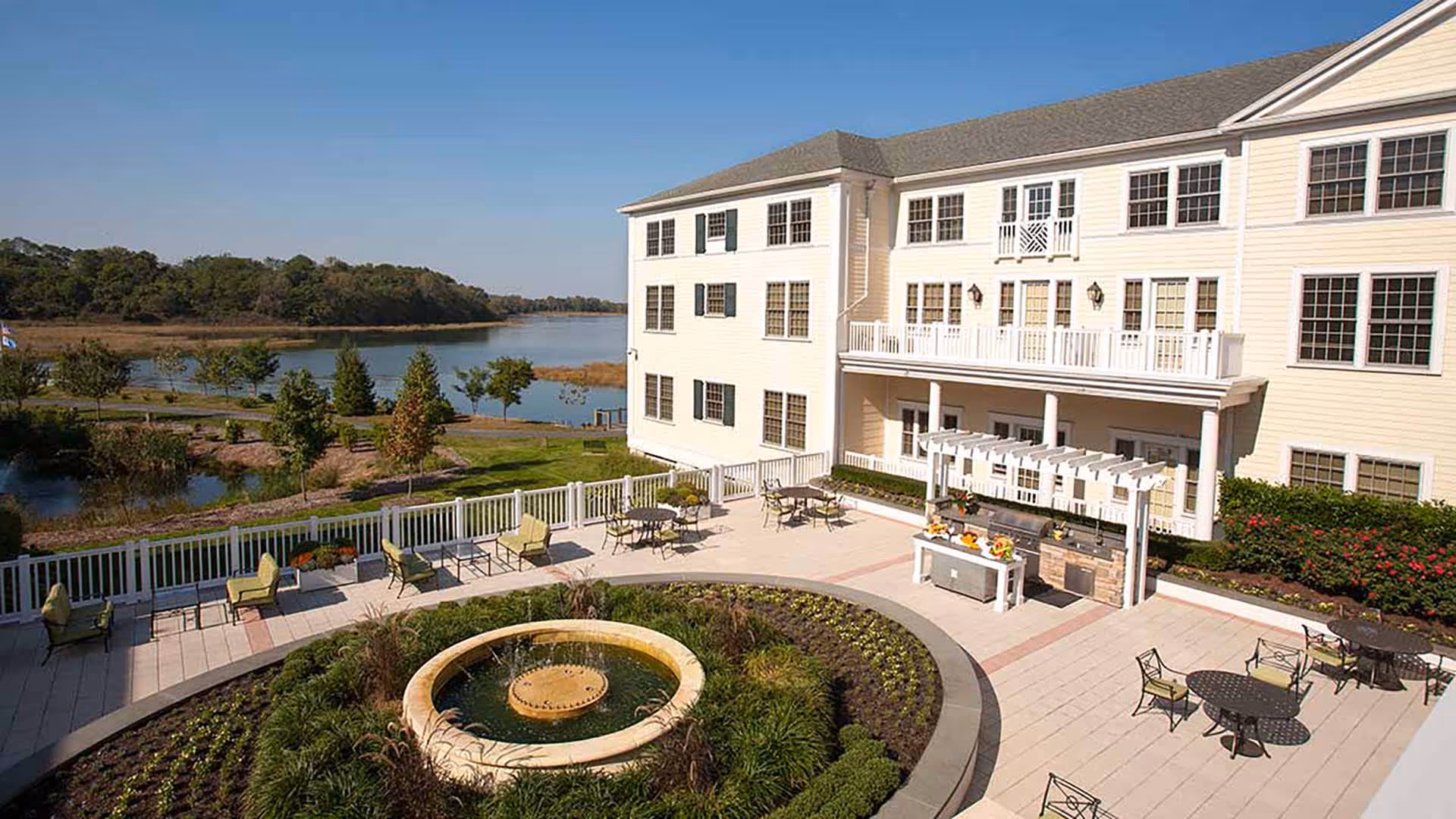 Outdoor patio area of a senior living facility with tables, chairs, a circular fountain surrounded by plants, and a built-in grill under a pergola. The building is a three-story light-colored structure with many windows. In the background, there is a scenic view of a river and trees under a clear blue sky.