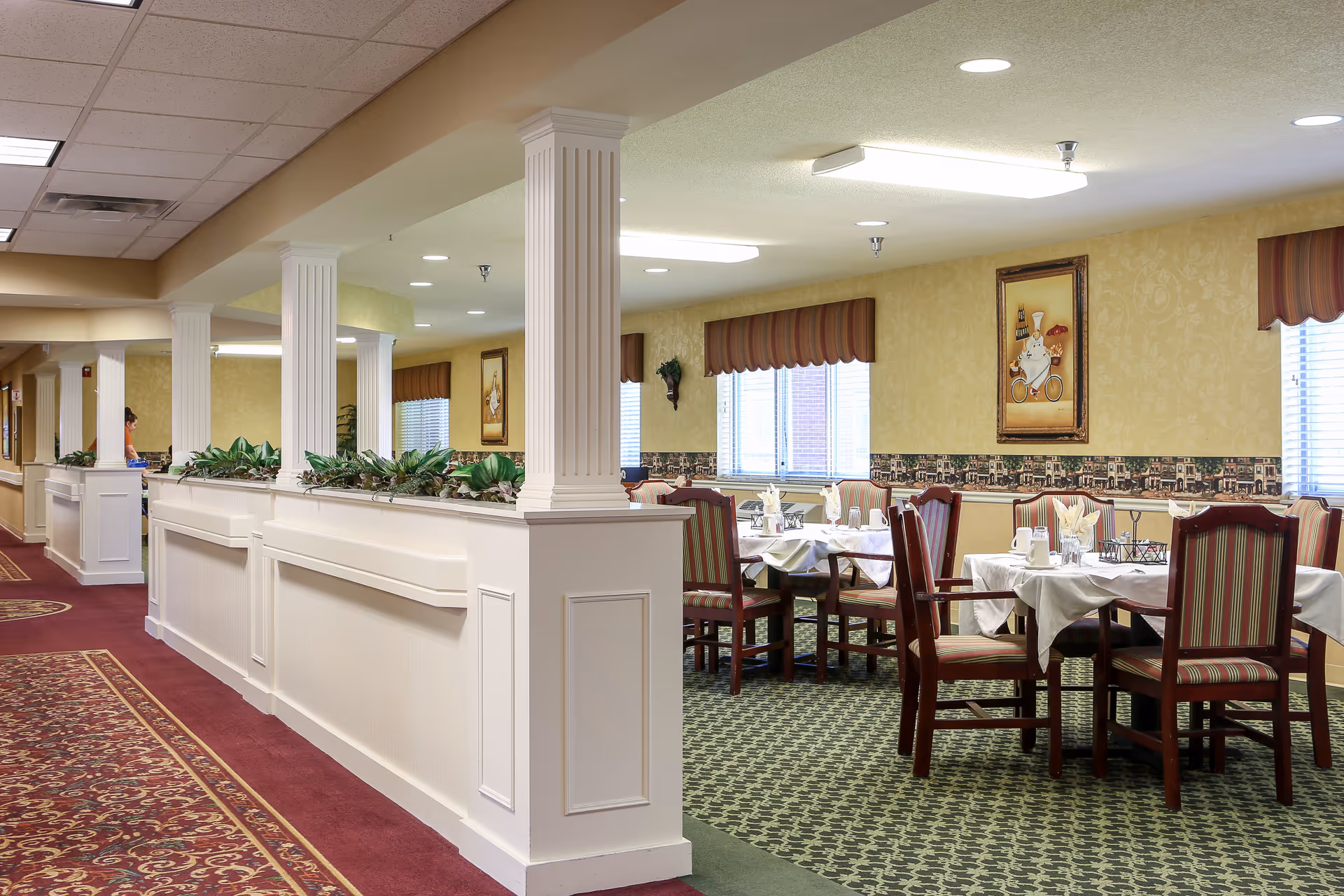 Dining room with multiple tables set with white tablecloths and striped chairs, separated from a carpeted corridor by white columns and planters.