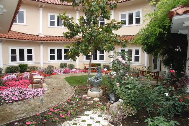 A courtyard garden area with a curved pathway, colorful flower beds, a small statue fountain, benches, and a two-story building with multiple windows surrounding the garden.