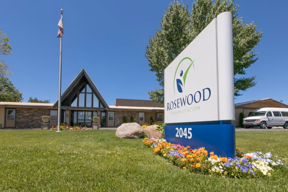 Exterior view of Rosewood Rehabilitation Center building with a large sign displaying the facility name and address number 2045, a well-maintained lawn with colorful flowers, an American flag on a flagpole, and a white van parked nearby under a clear blue sky.