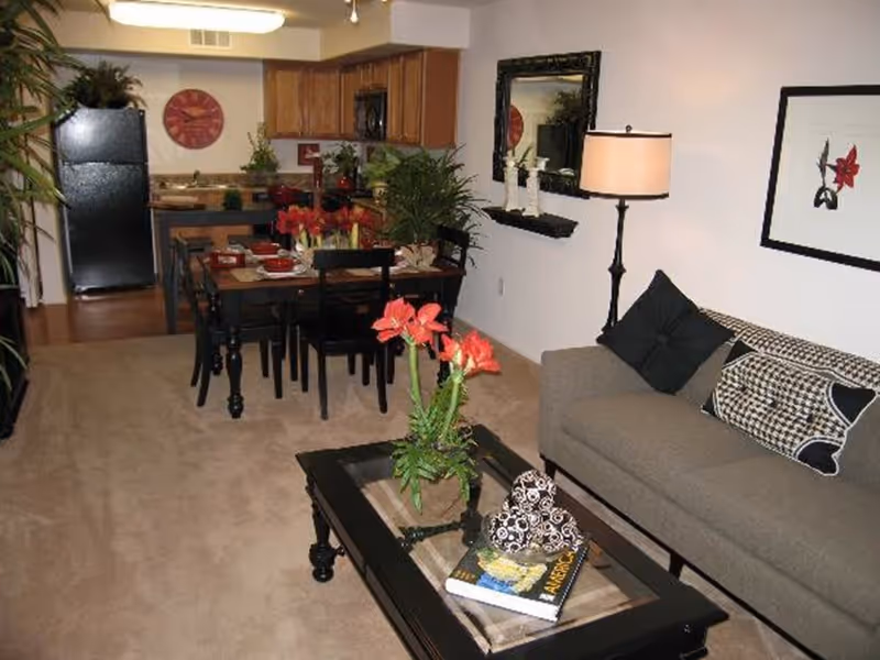 Interior view of a senior living facility showing a living room and kitchen area. The living room has a gray sofa with patterned cushions, a black coffee table with a flower vase and books, a floor lamp, and a wall mirror. The dining area has a black dining table set with red flowers and tableware. The kitchen in the background has wooden cabinets, a black refrigerator, and a red wall clock.