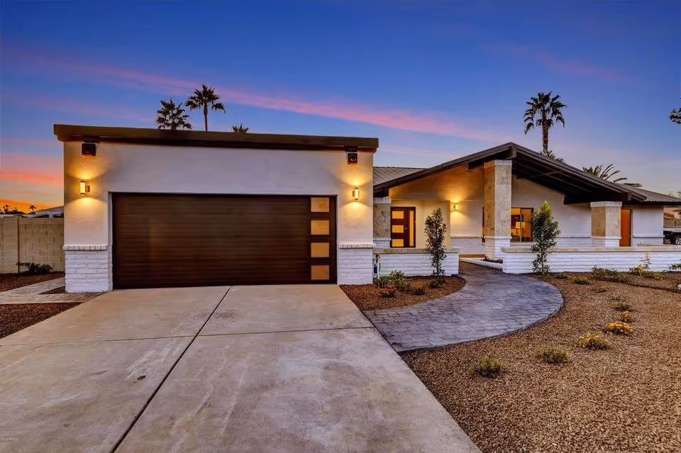 Front exterior of a modern single-story home with a two-car garage, lit porch, curved walkway, and palm trees at sunset.