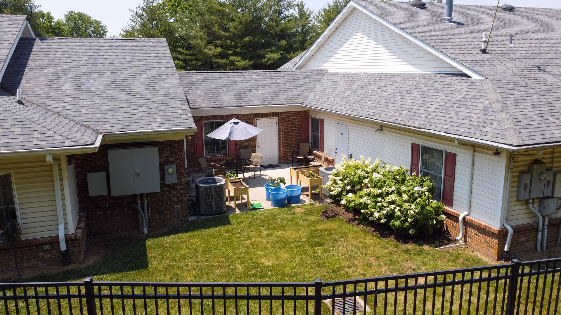 A fenced backyard area of a residential building with a small patio. The patio has outdoor chairs, a table with an umbrella, and several blue planters. There are flowering bushes along the side of the building, and the lawn is well-maintained. The building has a combination of brick and white siding with red shutters on the windows.