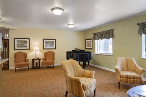 A cozy living room area with two striped armchairs in the foreground and two brown upholstered armchairs with a small table and lamp between them against a beige wall. A black grand piano is positioned near two windows with valance curtains, and the room is carpeted with a patterned design.