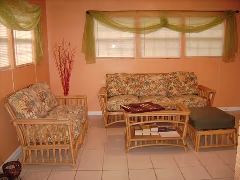 A cozy living room with peach-colored walls and white tiled floor. The room features a floral-patterned cushioned sofa set made of wicker, including a two-seater sofa, a single-seater chair, a glass-top coffee table with books underneath, and an ottoman with a green cushion. There are three windows with blinds and green sheer curtains above the sofa, and a decorative bundle of red sticks in the corner.