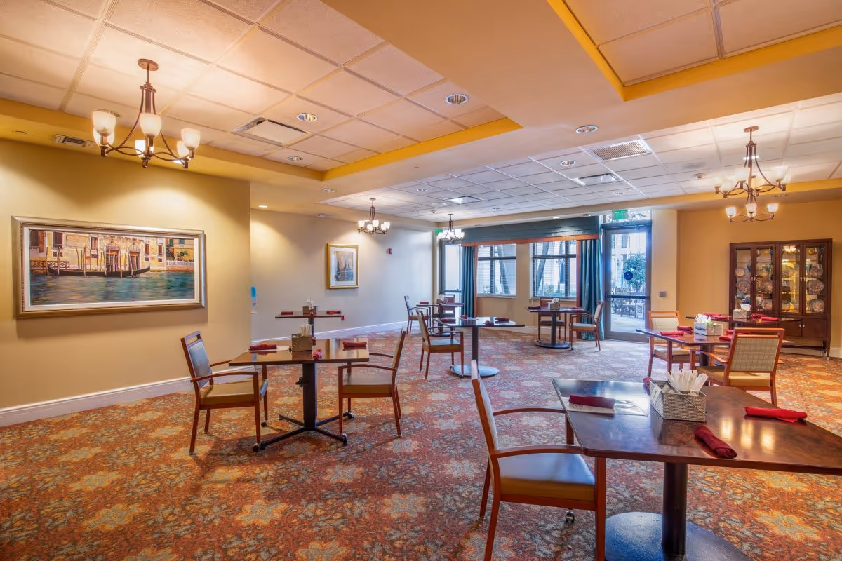 Spacious dining room with multiple small tables and chairs, chandeliers, wall art, and a display cabinet.