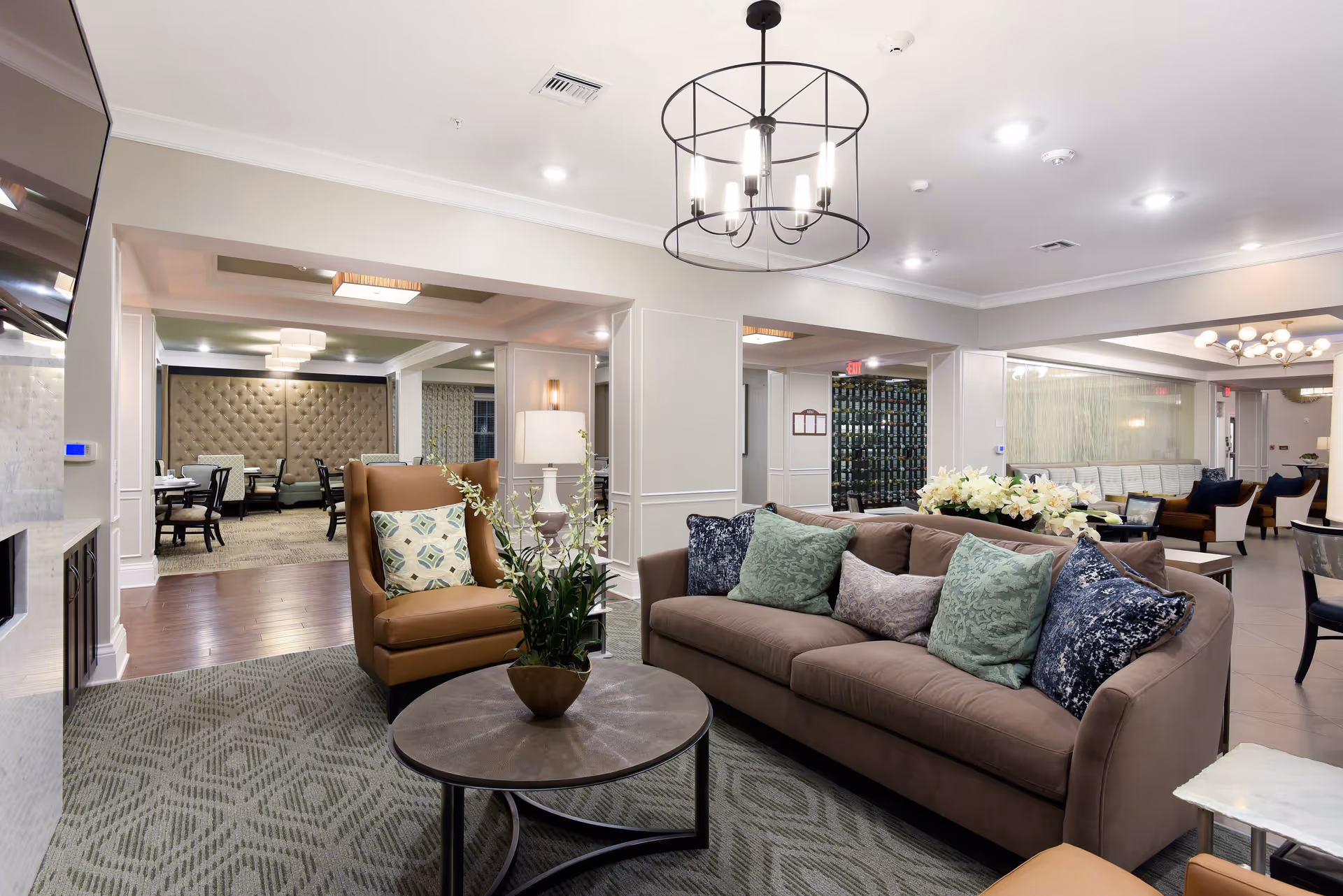 A modern and comfortable senior living facility common area featuring a brown sofa with multiple decorative pillows, a brown armchair with a patterned cushion, a round coffee table with a potted plant, and a stylish chandelier overhead. In the background, there is a dining area with tables and chairs, soft lighting, and elegant decor.
