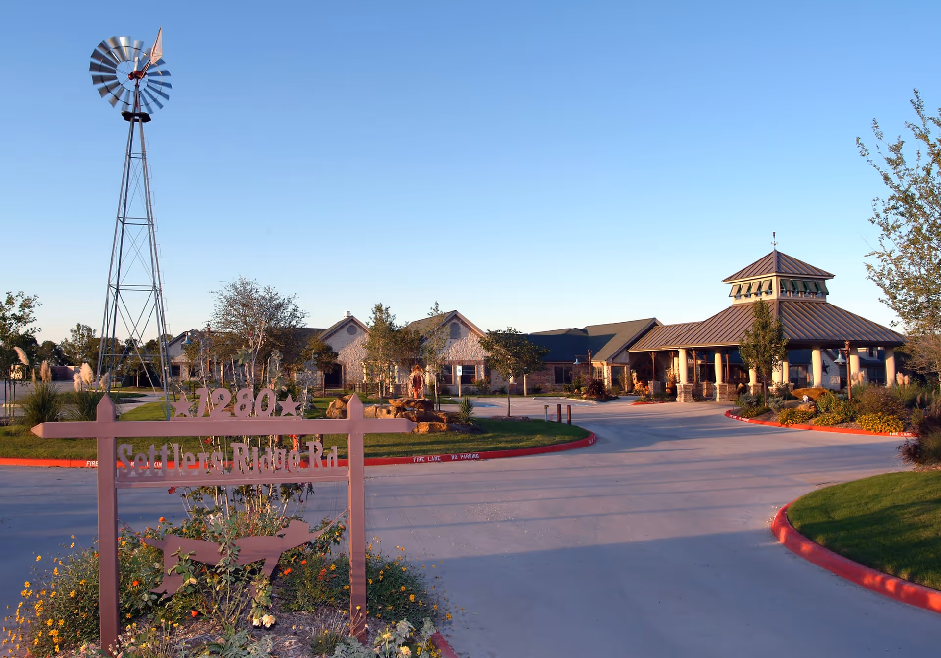 Exterior view of Settlers Ridge Care Center featuring a large driveway, landscaped garden with flowers and trees, a tall windmill structure, and a sign with the address 1280 Settlers Ridge Rd. The building has a rustic design with a covered entrance and a cupola on the roof under a clear blue sky.