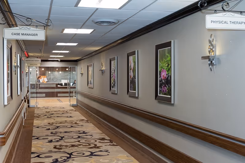 A well-lit hallway in St. James Rehabilitation & Healthcare Center with framed floral artwork on the walls, a patterned carpet, and signs indicating directions to Case Manager and Physical Therapy areas. At the end of the hallway, there is a reception desk with a person seated behind it.