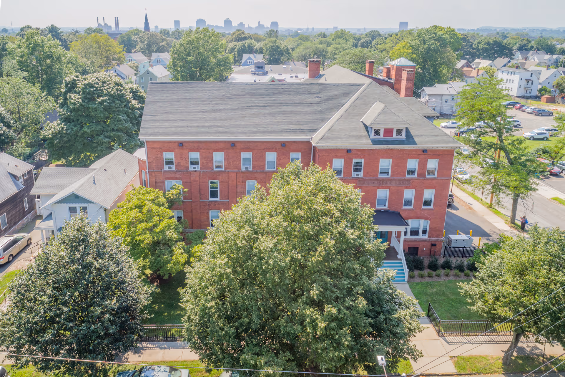 Aerial view of a three-story red-brick building surrounded by trees and nearby houses.