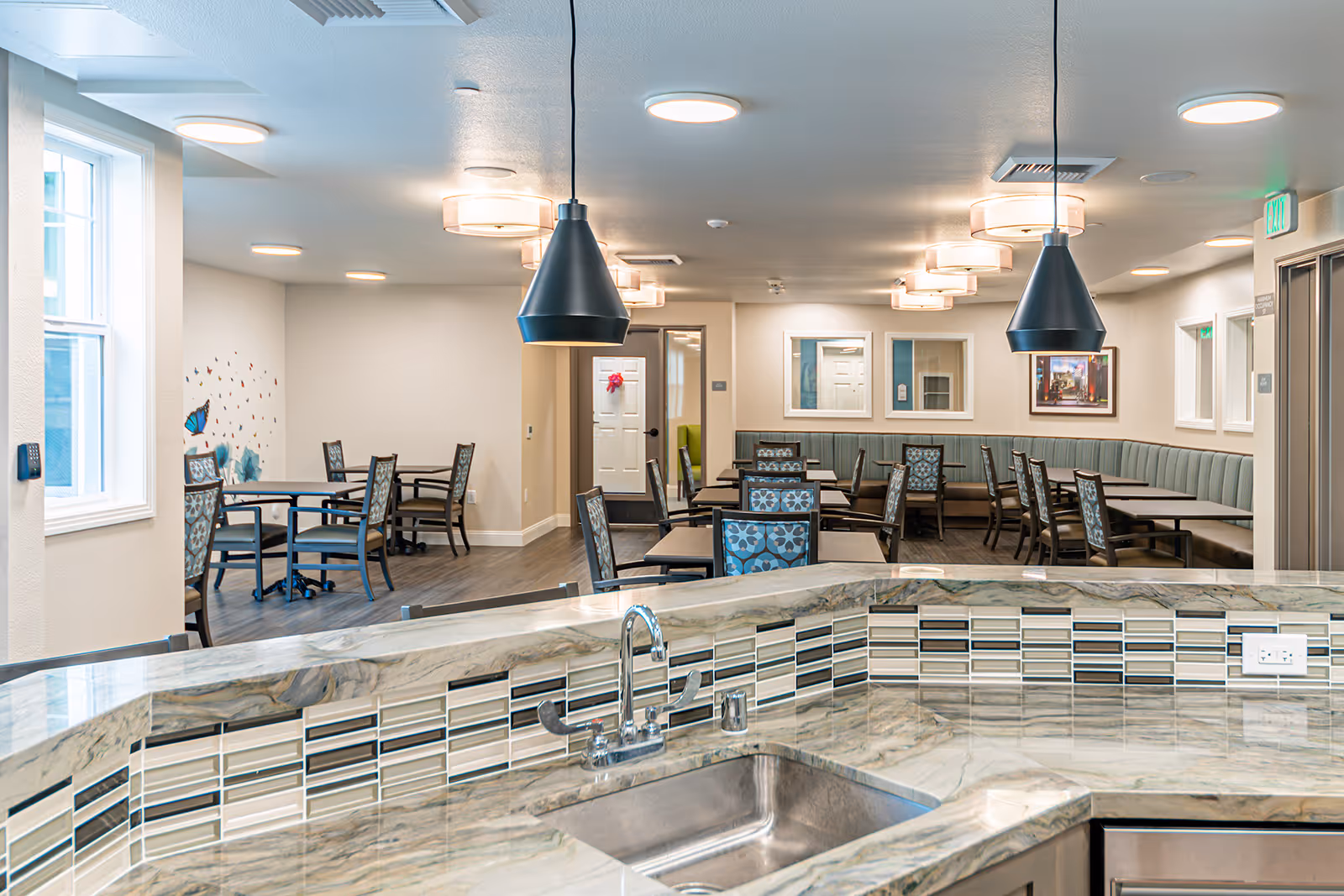 Interior view of a senior living facility dining area with multiple tables and chairs arranged neatly. The foreground shows a marble countertop with a sink and modern tile backsplash. The room is well-lit with ceiling lights and pendant lamps, and there are windows and wall decorations including butterfly decals.