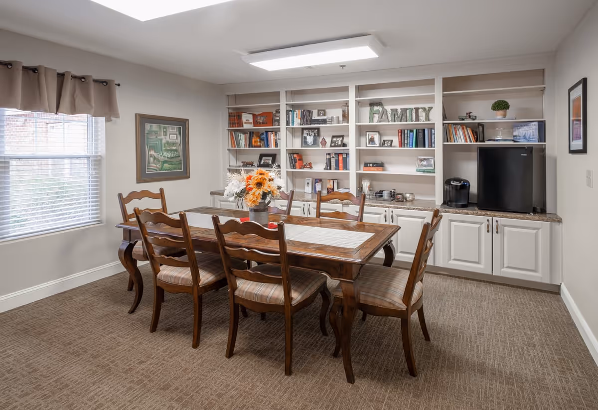 A cozy dining room with a wooden table and six matching chairs with cushioned seats. The table has a centerpiece of flowers. Behind the table is a built-in white shelving unit filled with books, framed photos, decorative items, and a small black refrigerator with a coffee maker on the countertop. A window with beige curtains allows natural light into the room, and a framed picture hangs on the wall.