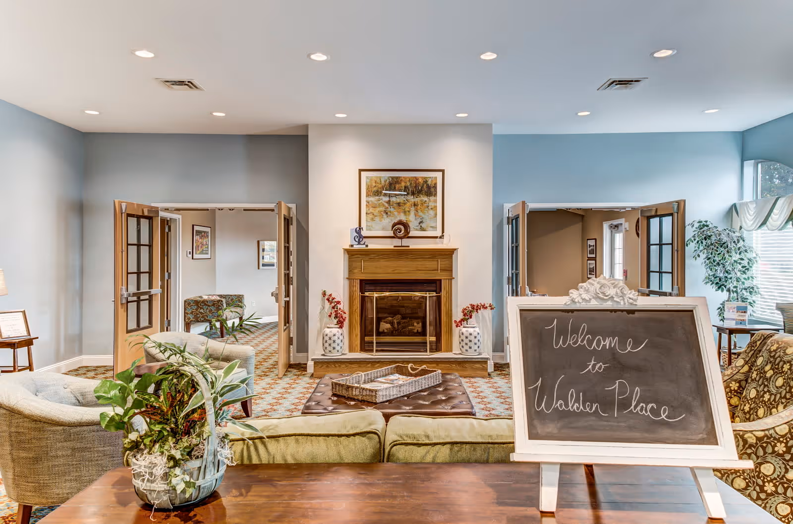 A cozy living room area in Walden Place featuring a fireplace with a framed painting above it, two open double doors on either side, comfortable armchairs, a coffee table with a tray, and a chalkboard sign that reads 'Welcome to Walden Place'. There are plants and soft lighting creating a warm and inviting atmosphere.