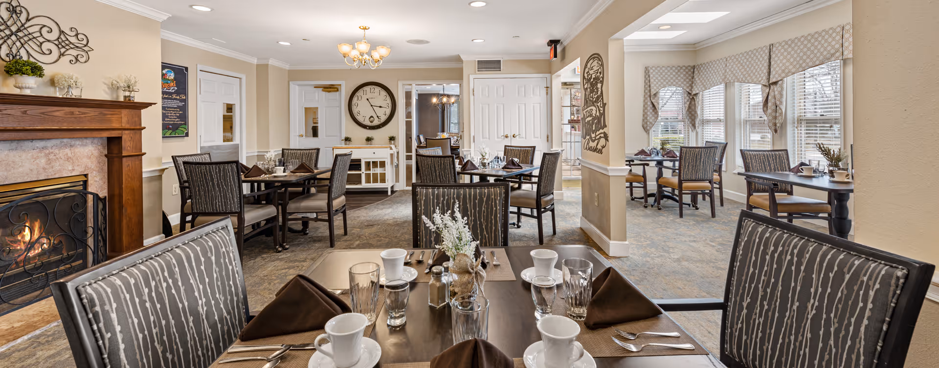 Dining room with set tables, upholstered chairs, a fireplace, and large windows in a senior living facility.