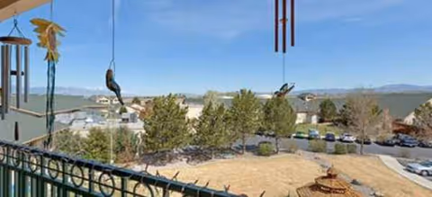 Balcony railing with wind chimes overlooks trees, a parking lot, and distant mountains.