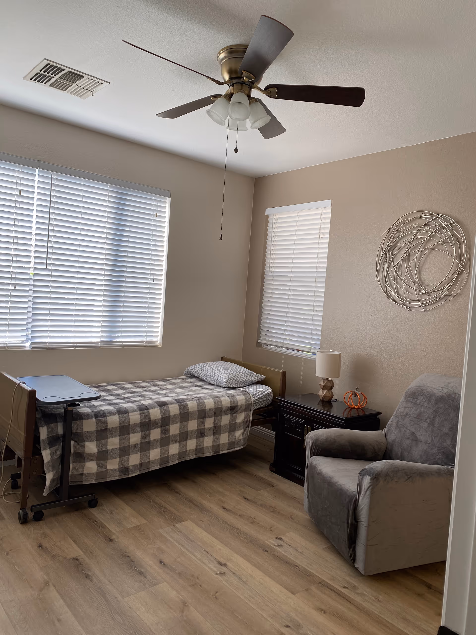 A small bedroom with a single bed covered in a checkered blanket and a pillow, a wooden nightstand with a lamp and a small decorative pumpkin, a gray upholstered armchair, two windows with white blinds, a ceiling fan with lights, and a circular metal wall decoration.