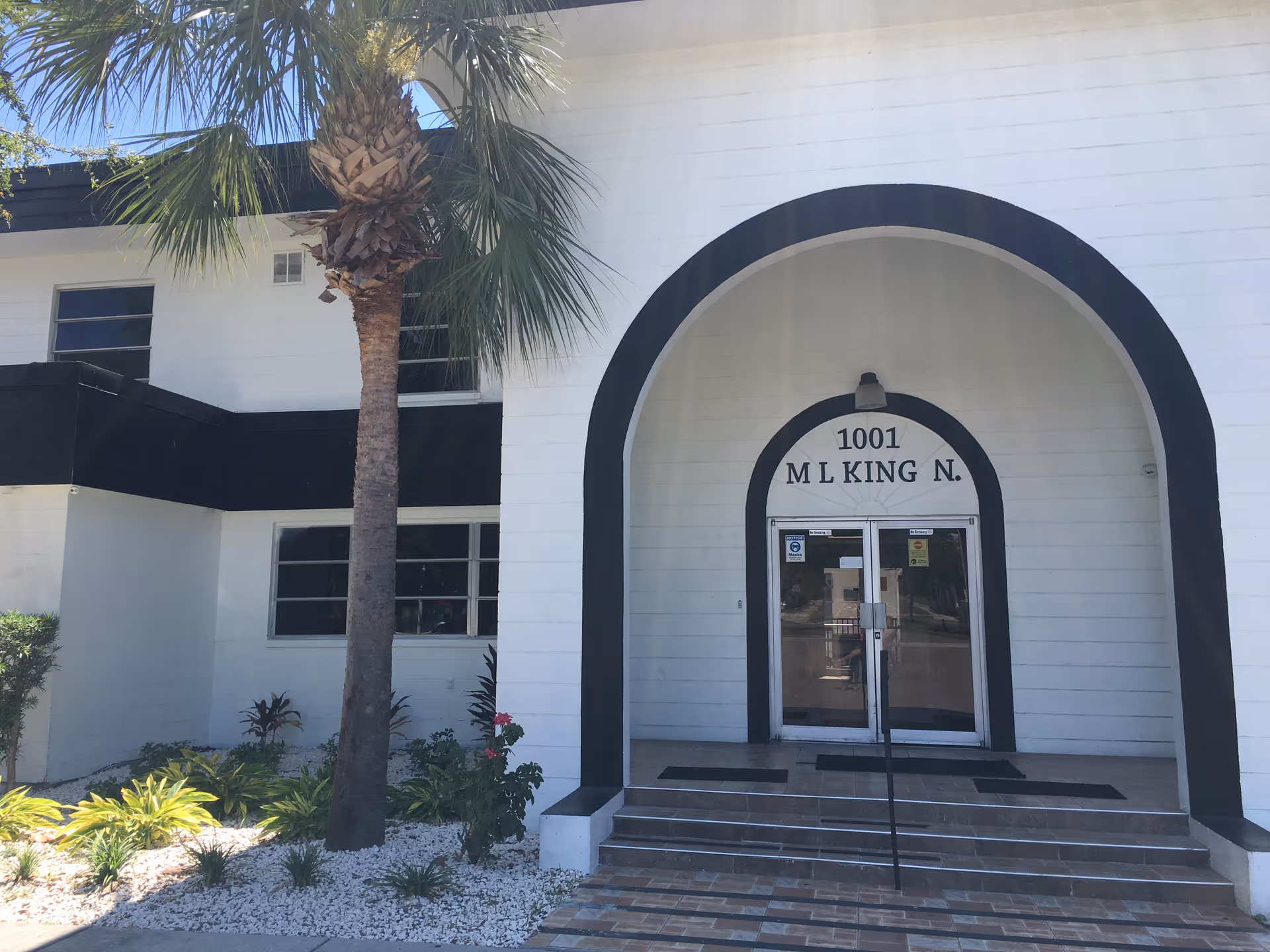 Entrance of a white building with black trim featuring an arched doorway. The address 1001 M L King N. is displayed above double glass doors. There is a palm tree and landscaped plants with white rocks in front of the building.