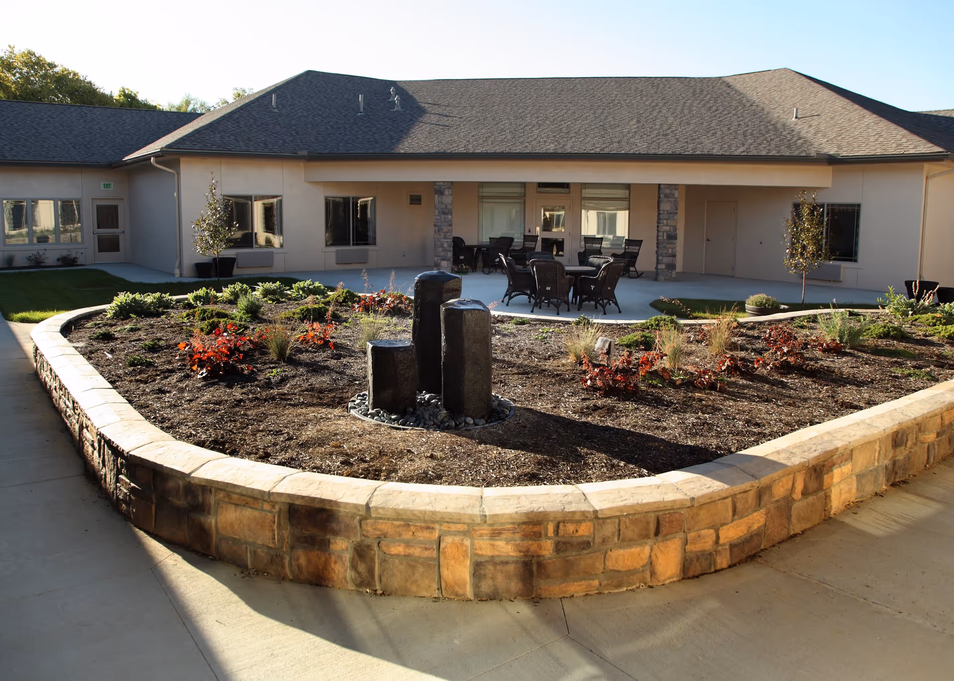 Outdoor courtyard area at Windsor Heights Assisted Living and Memory Care featuring a raised stone planter with various plants and a small water fountain in the center. Surrounding the planter is a concrete walkway, and in the background, there is a building with a covered patio area furnished with tables and chairs.
