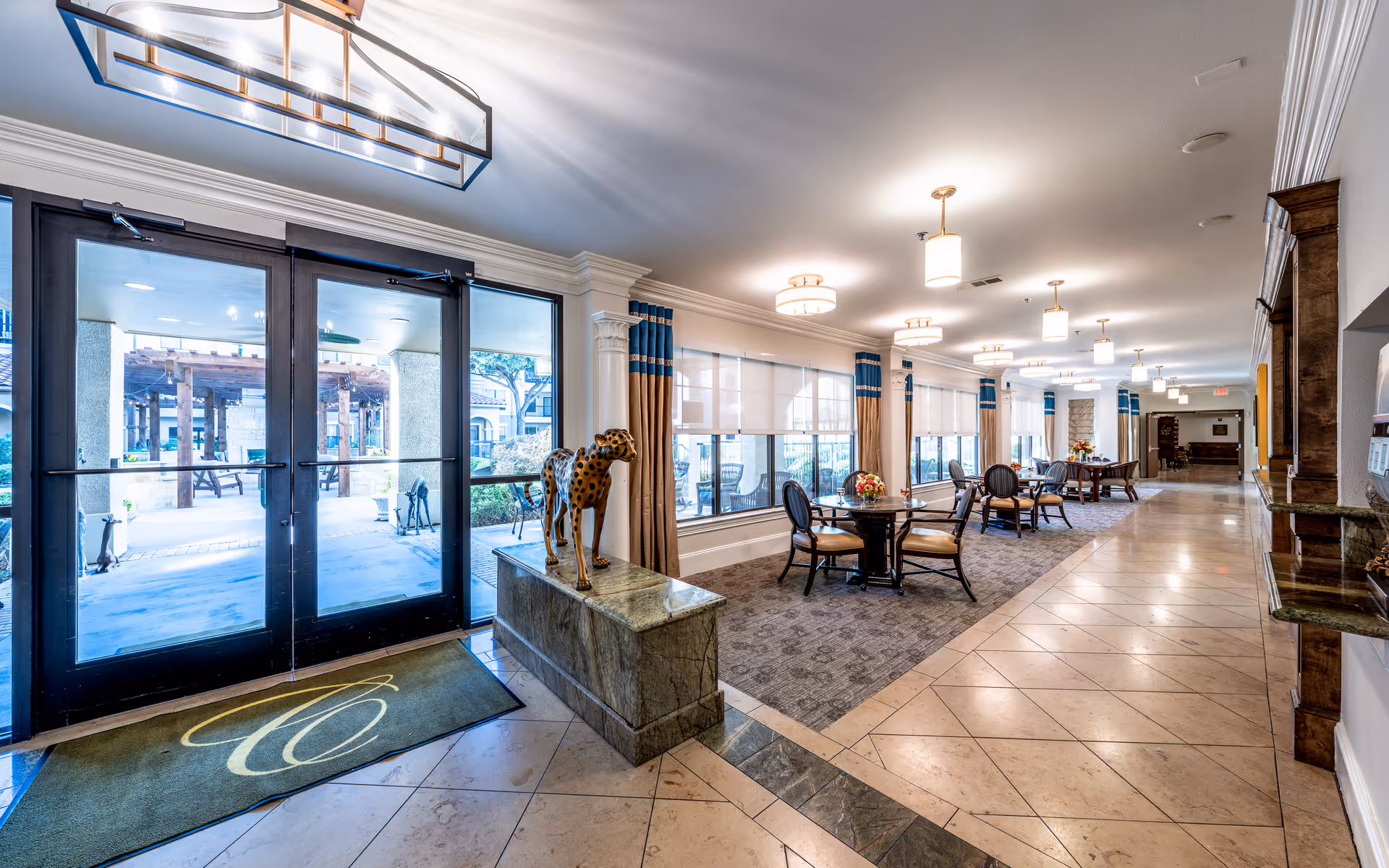 Bright and spacious interior hallway of a senior living facility with large windows covered by white blinds and blue curtains. Several round tables with chairs are arranged along the carpeted area near the windows, each table decorated with a flower arrangement. A decorative cheetah statue stands on a stone pedestal near glass double doors leading outside to a patio area with pergolas and benches. The floor is tiled, and the ceiling has multiple modern light fixtures.