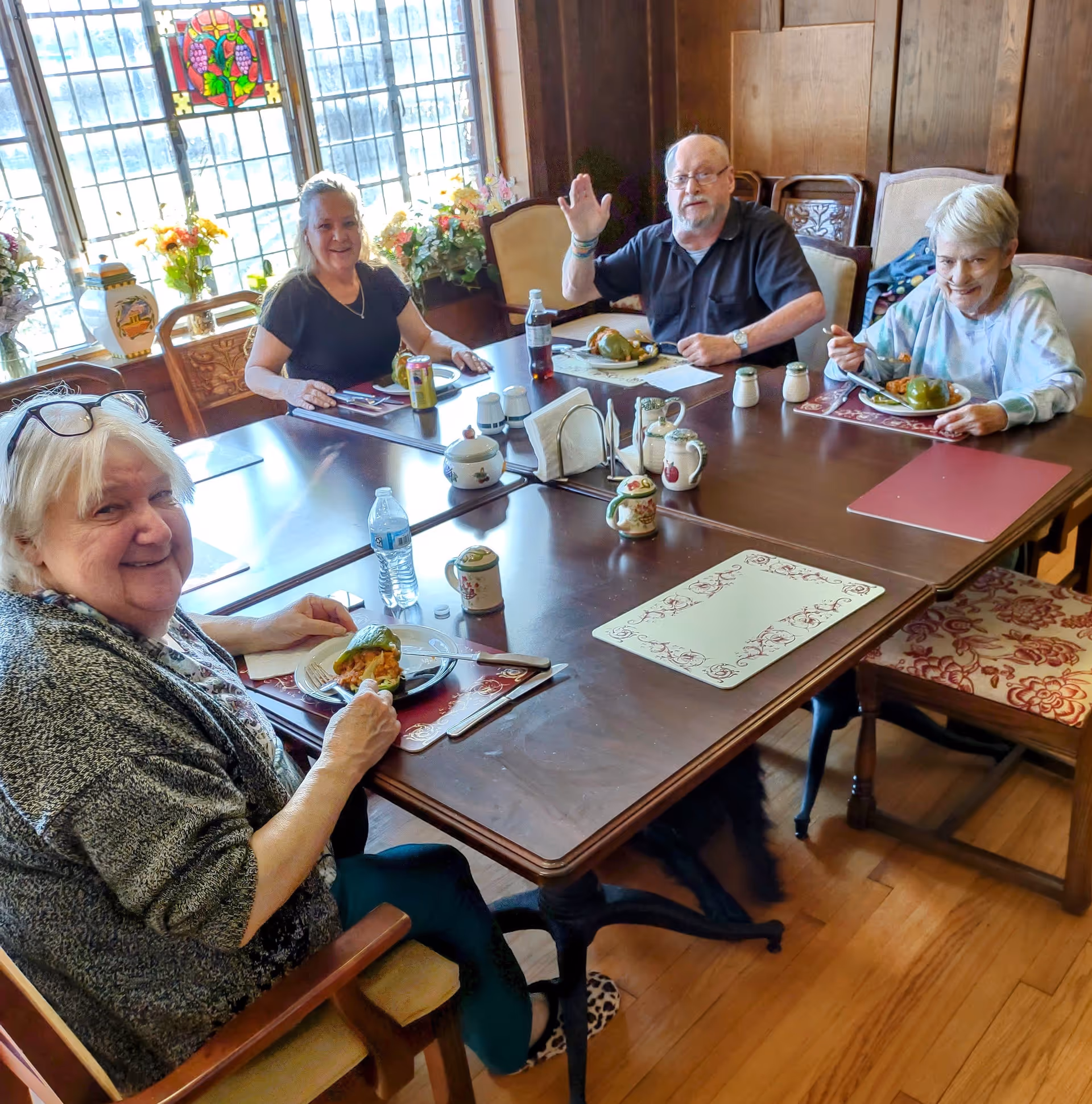 Four elderly people sitting around a wooden dining table enjoying a meal together in a room with stained glass windows and floral decorations. One man is waving at the camera while the others smile.
