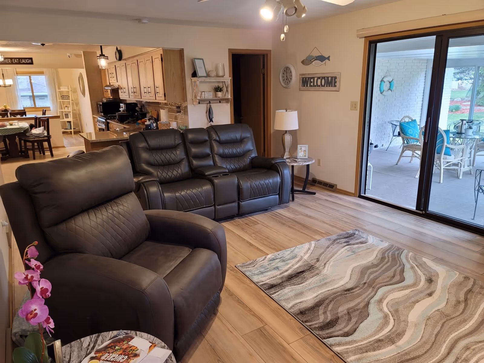 A cozy living room with a dark leather armchair and a matching leather loveseat. The room has light wood flooring and a patterned area rug. To the right, there is a sliding glass door leading to a covered patio with outdoor seating. In the background, a kitchen and dining area are visible with wooden cabinets and a dining table. The wall has a clock, a welcome sign, and decorative fish artwork.