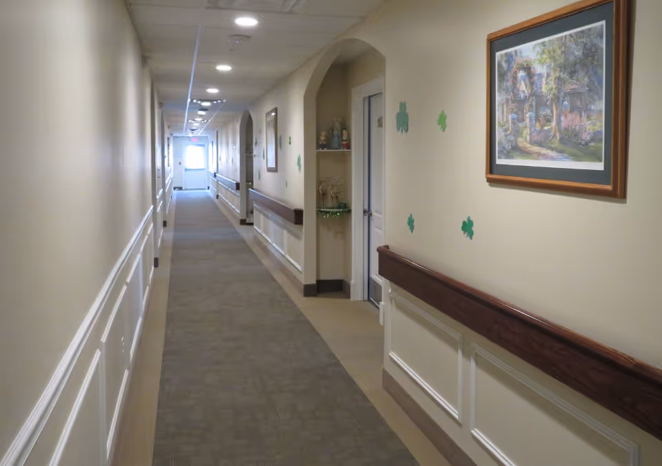Long carpeted hallway inside an assisted living facility with handrails, framed art, and decorative clover decals on the walls.