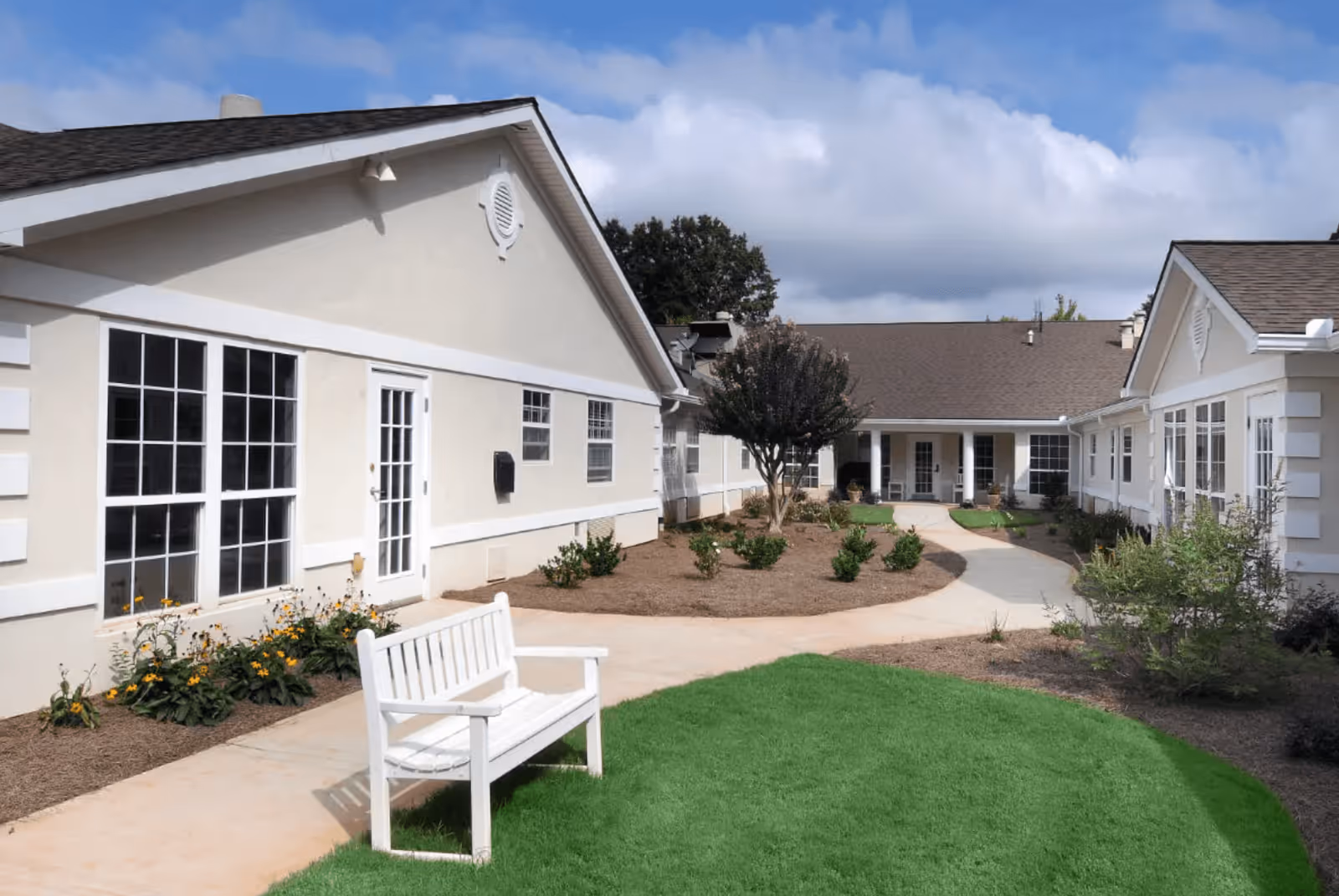 Outdoor courtyard area of a senior living facility with beige buildings surrounding a landscaped garden. There is a white bench on a concrete path, green grass, small bushes, and a tree in the center. The sky is partly cloudy.