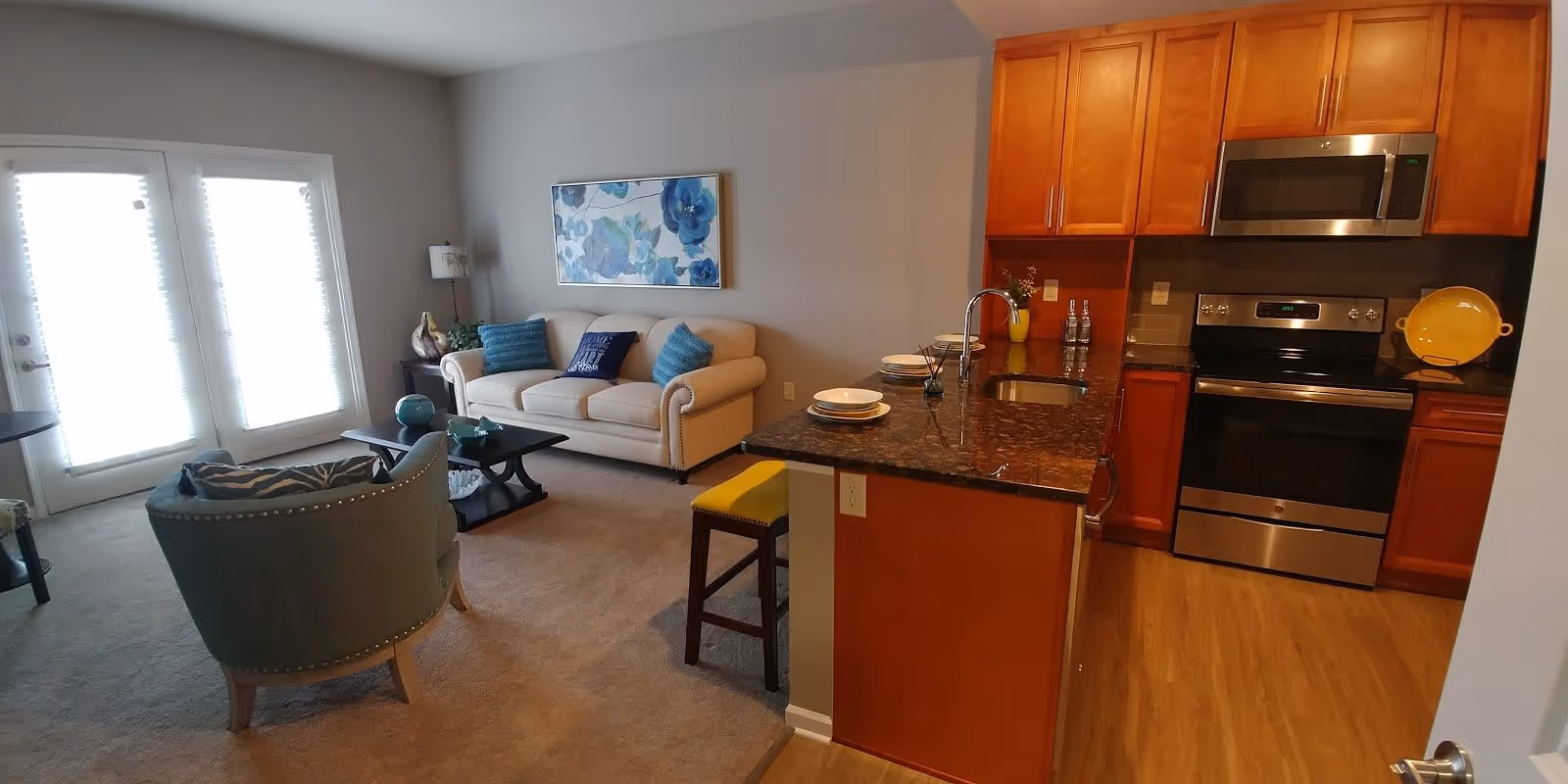 A cozy living room and kitchen area in an apartment. The living room features a beige sofa with blue and patterned throw pillows, a green armchair, a coffee table, and a side table with a lamp. There is a large floral painting on the wall above the sofa. The kitchen has wooden cabinets, a granite countertop with a sink, a stove, microwave, and a yellow stool at the counter. French doors with blinds allow natural light into the room.