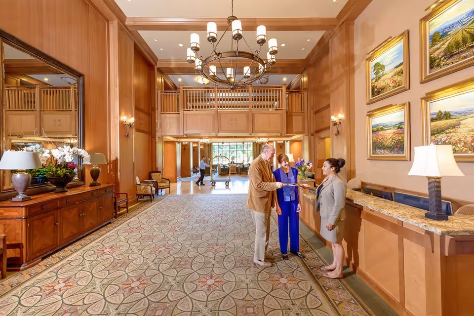 A spacious and elegant senior living facility lobby with a high ceiling, large chandelier, and wooden paneling. A man and a woman are interacting with a receptionist at the front desk. The lobby features a patterned carpet, a large mirror above a wooden console with lamps and flowers, and framed landscape paintings on the wall.