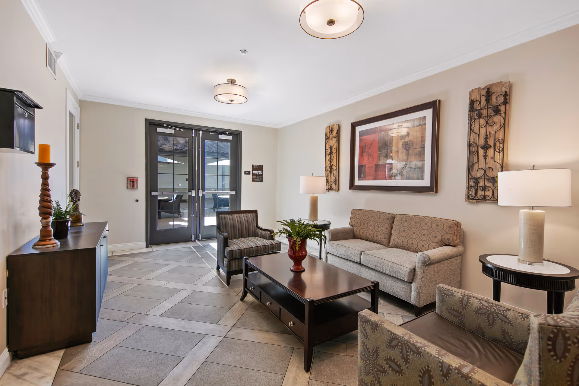 A well-lit living room area with a beige couch, two patterned armchairs, a dark wooden coffee table with a red vase and green plant, two side tables with lamps, decorative wall art, and double glass doors leading outside.