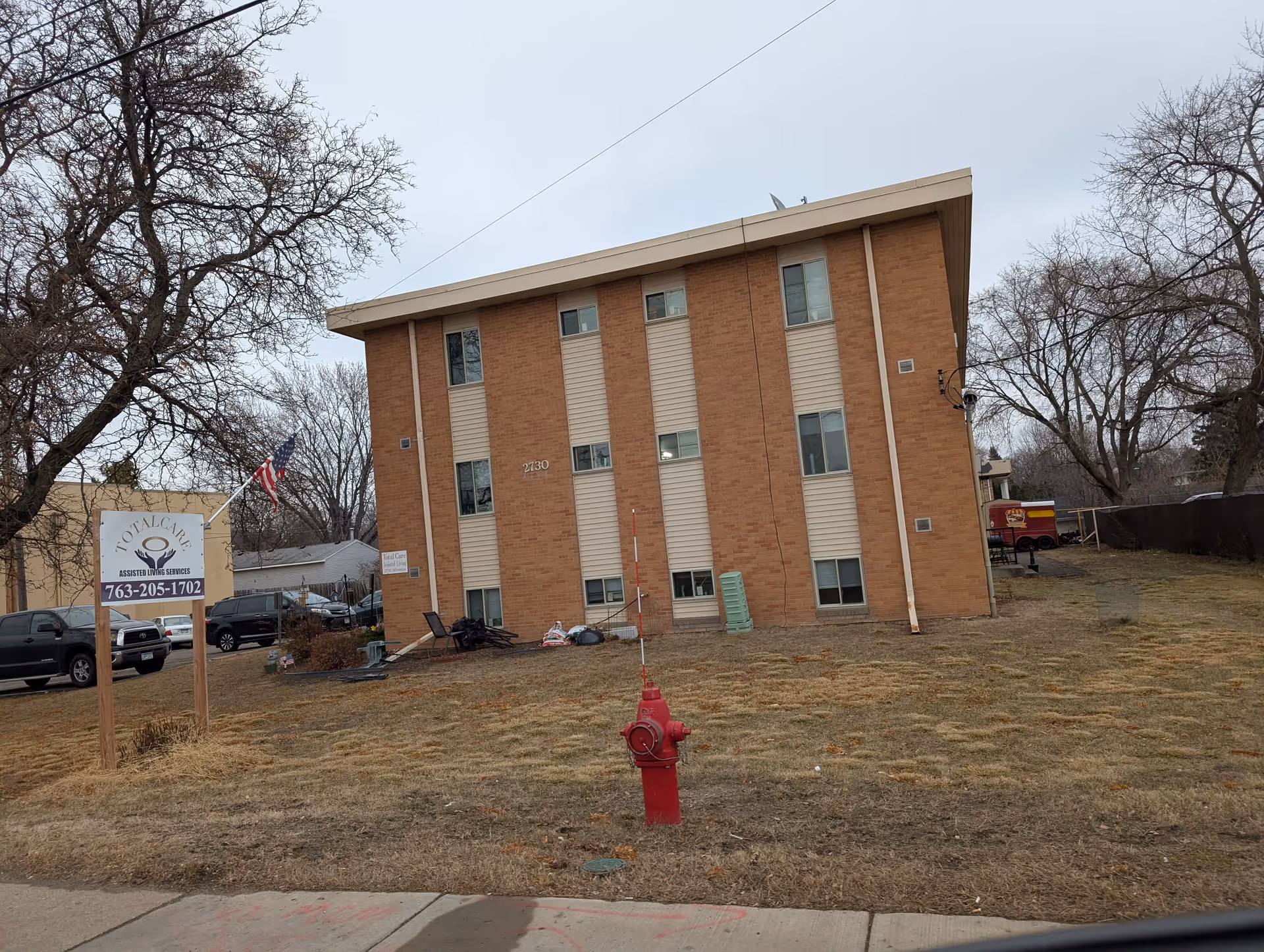 Exterior view of a three-story brick building with beige vertical panels and multiple windows. In front of the building is a red fire hydrant on a patch of dry grass. To the left, there is a sign for Total Care Assisted Living Services with a phone number and an American flag. Several parked cars and leafless trees are visible around the building under a cloudy sky.