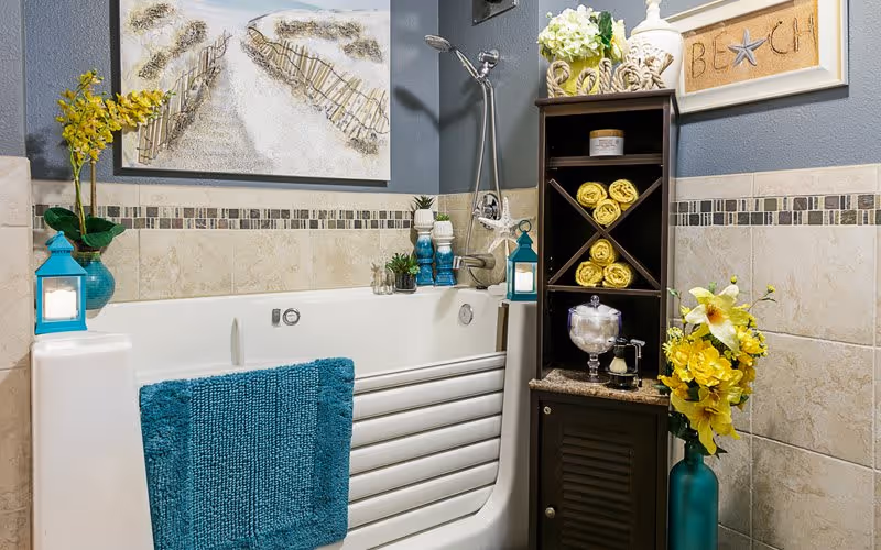 A bathroom featuring a white walk-in bathtub with a blue towel hanging on the side. The walls are tiled with beige tiles and a decorative mosaic border. There is a dark wooden shelving unit next to the tub holding rolled yellow towels, a glass jar, and decorative items. The room is decorated with yellow flowers in blue vases and beach-themed artwork on the walls.