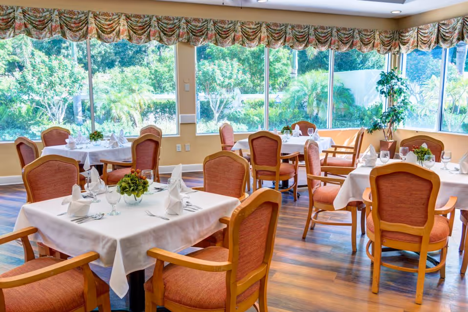 Sunlit dining room with neatly set tables and upholstered chairs overlooking a garden through large windows.