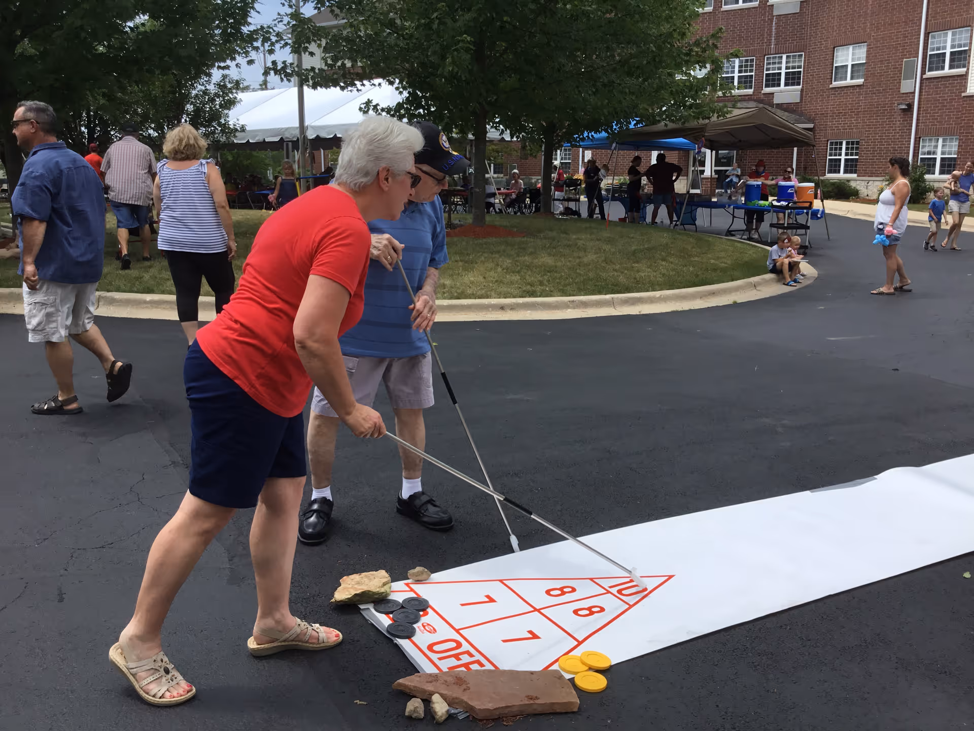 Two elderly people playing shuffleboard on a large outdoor shuffleboard court mat placed on an asphalt surface. Other people are walking and sitting under tents in the background near a brick building with multiple windows. Trees and grass are also visible.