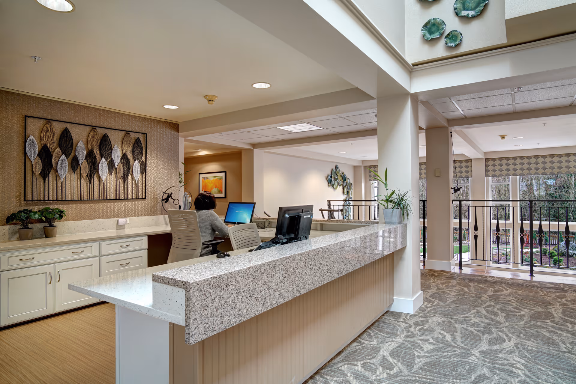 Reception area in a senior living facility with a granite countertop desk, a person seated working on a computer, decorative wall art featuring leaves, potted plants, and large windows overlooking a garden outside.