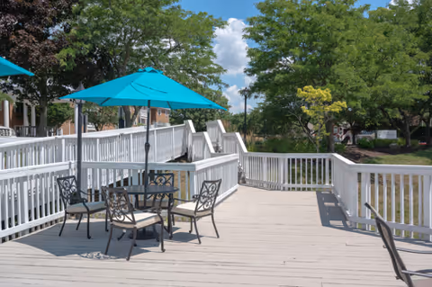 Outdoor wooden deck area with a black metal table and four chairs, shaded by a blue umbrella. The deck is surrounded by white railings and overlooks green trees and a grassy area under a partly cloudy sky.