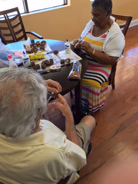 Two elderly individuals sitting at a round table engaged in a craft activity involving small objects and glue. The woman is wearing a colorful striped dress and white cardigan, while the man is wearing a light yellow shirt and shorts. The room has wooden flooring and large windows letting in natural light.