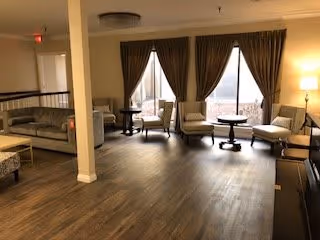 A spacious living room area with wooden flooring, featuring multiple upholstered chairs and sofas arranged around small round tables. Large windows with brown curtains allow natural light to enter the room. A floor lamp is visible on the right side, and a white pillar stands near the center of the room.